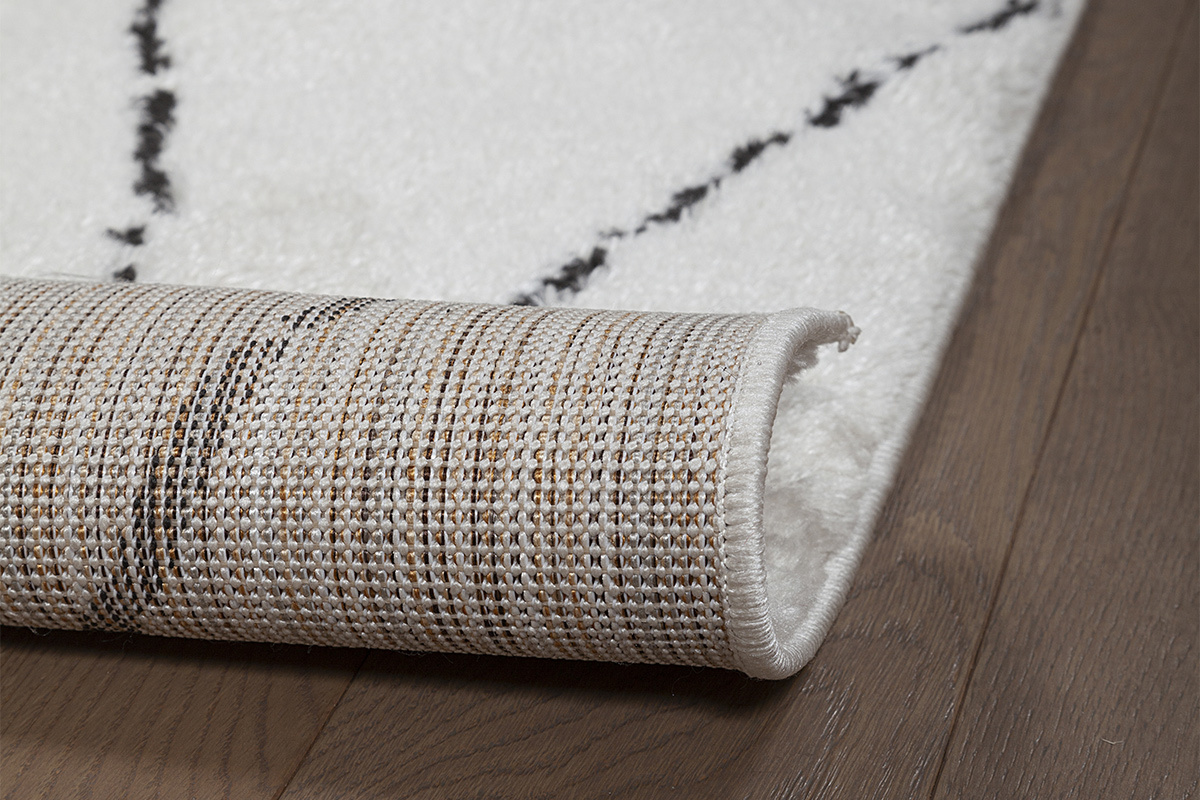 Primer plano de la alfombra SILAN con motivos negros y textura de tela, parcialmente enrollada.