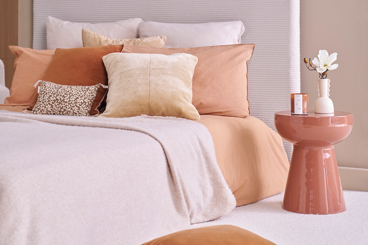 Mesa auxiliar redonda de metal esmaltado terracota al lado de una cama con cojines beige y terracota.