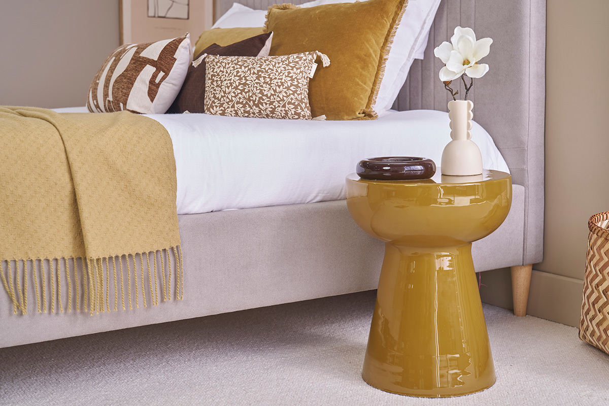 Mesa auxiliar redonda de metal esmaltado en bronce, cerca de una cama con cojines decorativos.