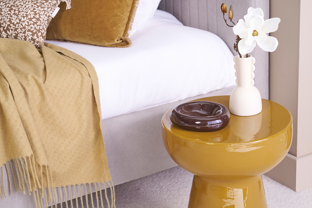 Mesa auxiliar MOGOU redonda bronce con jarr�n blanco y cenicero marr�n, al lado de una cama.