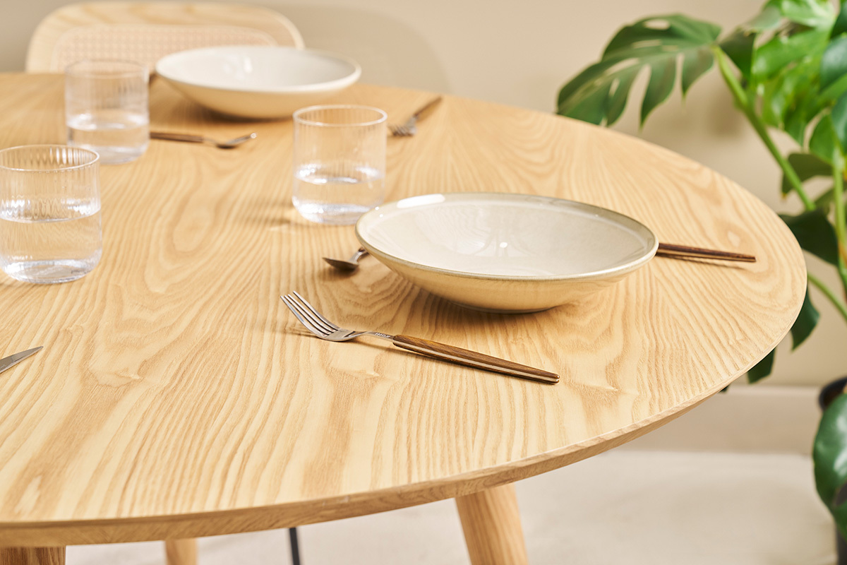 Mesa redonda de roble claro de 120 cm con vajilla y cubiertos, vista desde arriba.