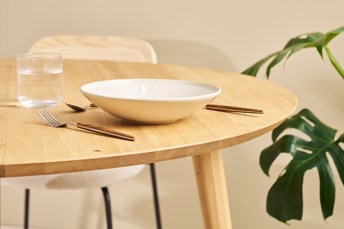 Mesa redonda de roble claro con vajilla y planta al fondo, vista cercana.