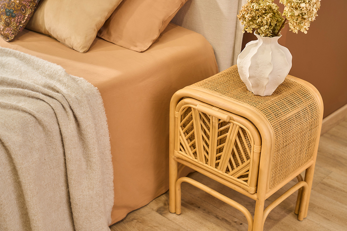 Mesita de noche de rat�n GALON, vista lateral, con jarr�n, al lado de una cama beige.