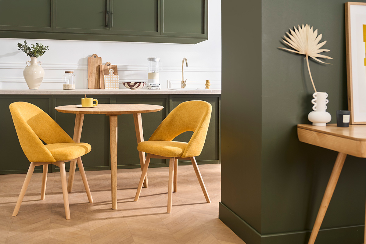Dos sillas escandinavas de tela amarilla mostaza y madera clara alrededor de una mesa redonda de madera.