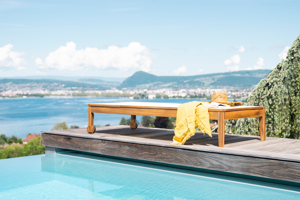 Tumbonas NAVEEN de madera maciza junto a una piscina, con una toalla amarilla y un sombrero de paja.