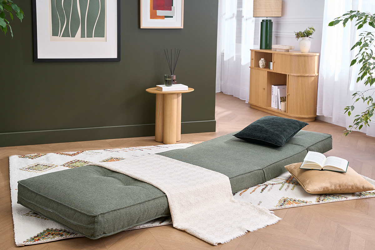 Sill�n convertible SALLY en tela verde caqui, desplegado, con cojines y manta sobre alfombra.