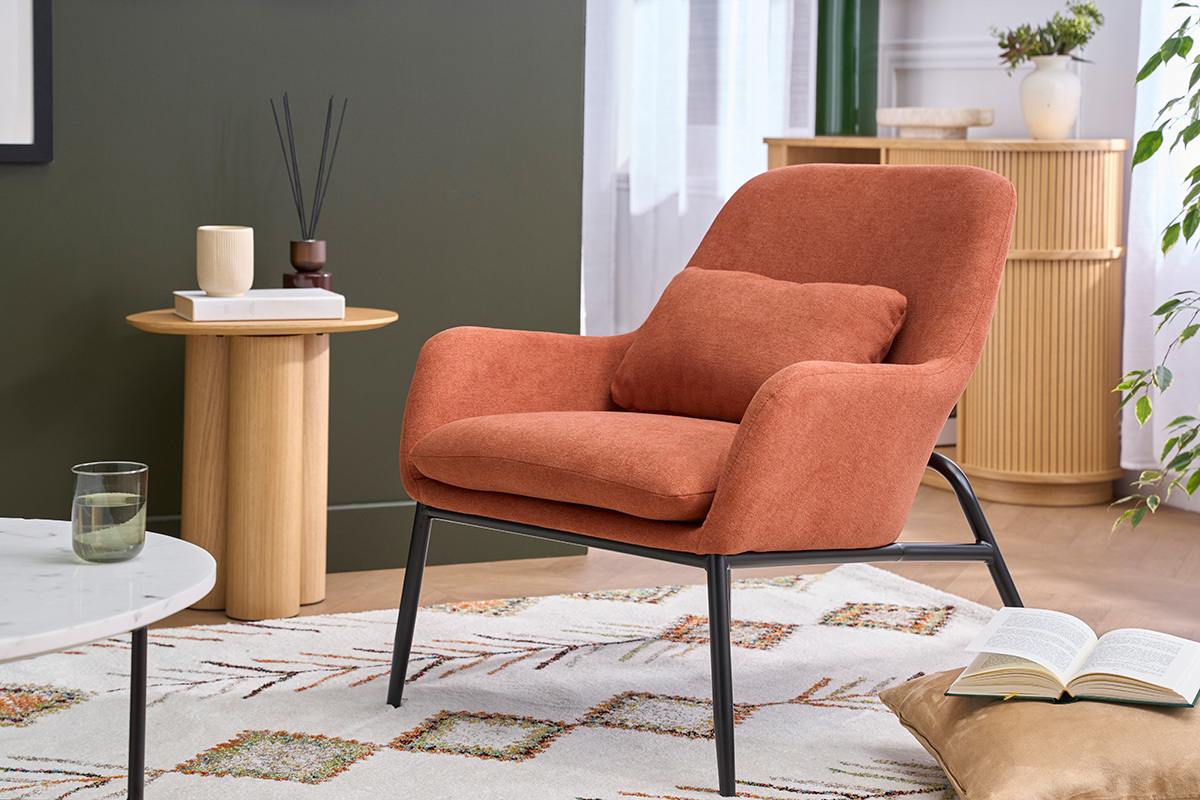 Sill�n de tela efecto terciopelo tierra quemada con patas de metal, �ngulo 3/4, al lado de una mesa de madera.