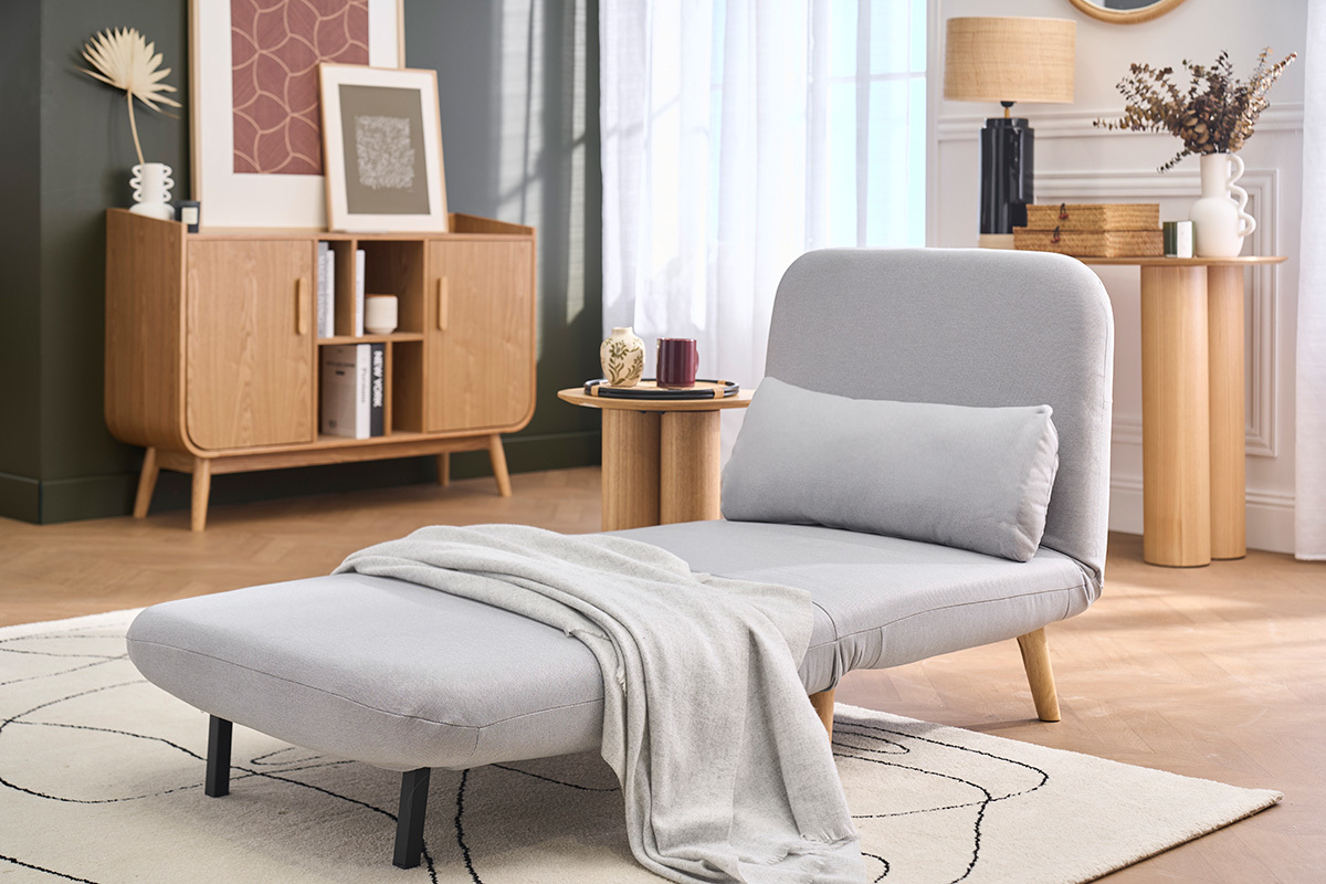 Sill�n convertible AMIKO gris claro en tela, vista 3/4, con manta, mesita auxiliar y decoraci�n escandinava.
