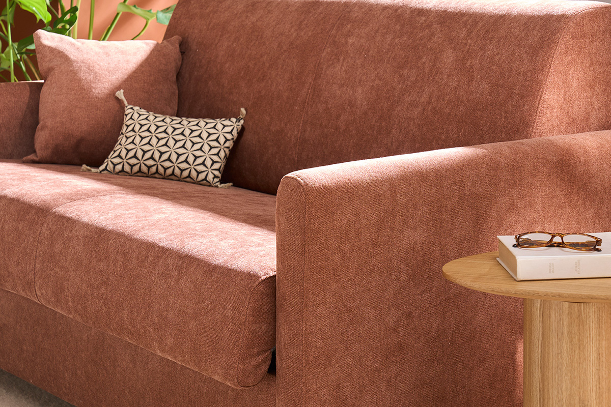 Sof convertible en terciopelo color tierra quemada, con brazo derecho y cojines con mesita de madera.