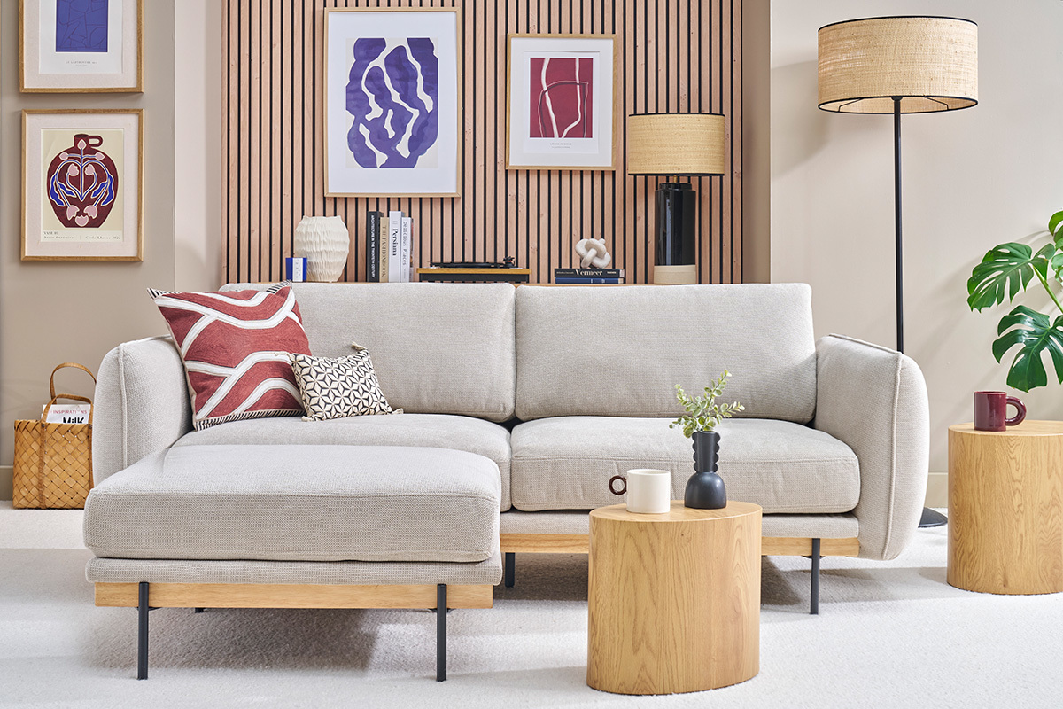 Sof� beige de 3 plazas con reposapi�s, vista frontal, rodeado de mesas de madera y decoraci�n mural moderna.