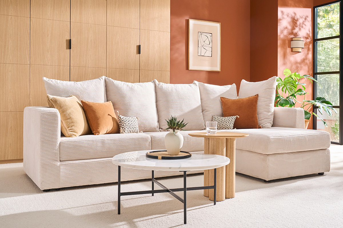 Sof esquinero convertible beige en terciopelo acanalado con cojines, mesa baja de mrmol, vista lateral.