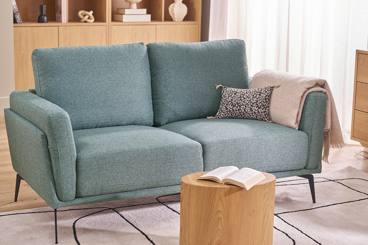 Sof� de dos plazas verde gris�ceo con patas negras, vista de 3/4 en una sala de estar.