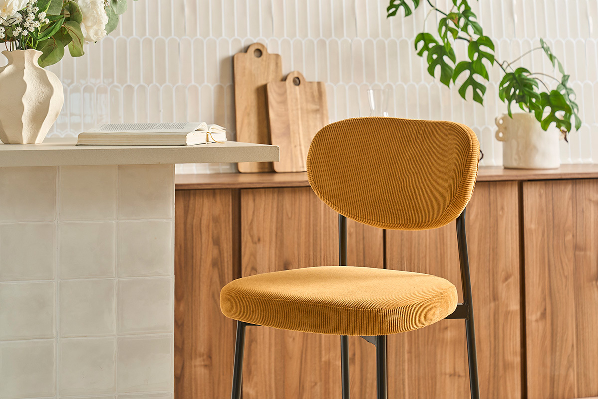 Taburete de bar fijo en tela de terciopelo de pana amarillo curry, vista lateral, con decoraci�n de fondo.
